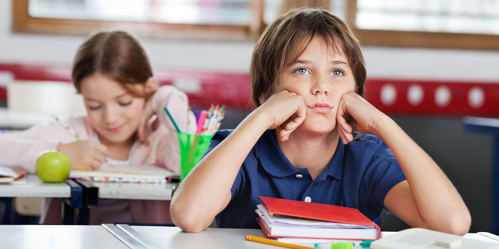 Stock image: Bored schoolboy looking away while sitting at desk with girl in background at classroom | By: Tyler Olson